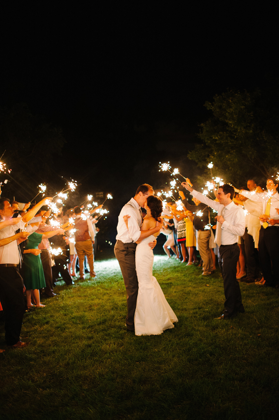 The bride and groom share one more kiss and then exit their wedding reception while guests hold sparklers at a backyard wedding by Bloomfield Hills wedding photographer Heather Jowett.