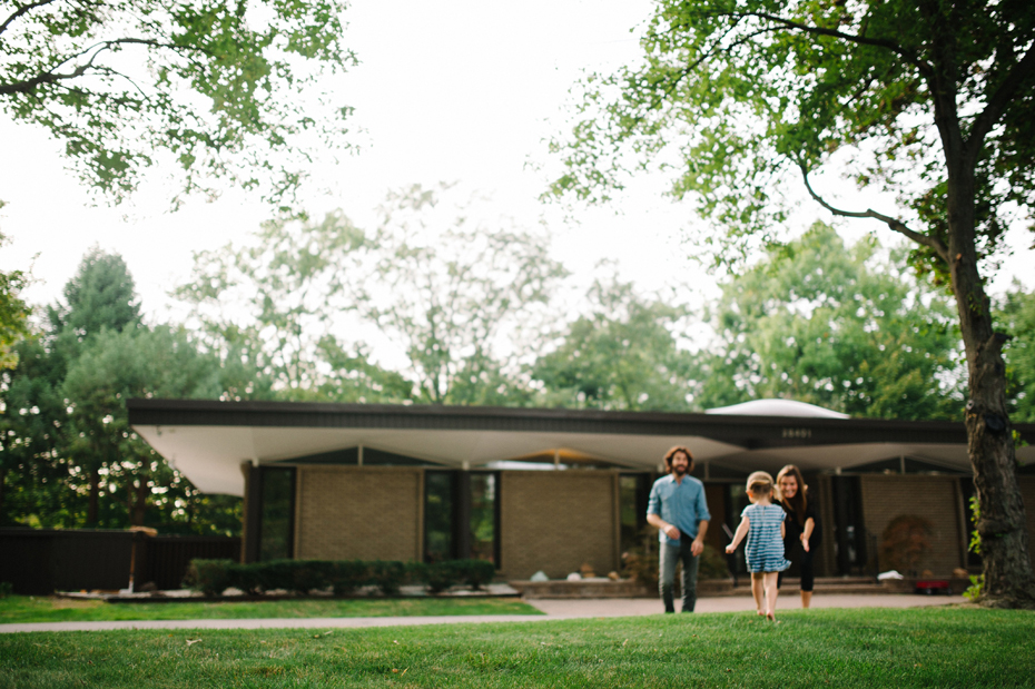 A two year old plays with her mother and father in the front yard of their mid-century home in Metro-Detroit during a photojournalistic family portrait session in Ferndale photographed by Ann Arbor Wedding Photographer, Heather Jowett.