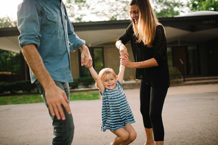 A two year old plays with her mother and father in the front yard of their mid-century home in Metro-Detroit during a photojournalistic family portrait session in Ferndale photographed by Ann Arbor Wedding Photographer, Heather Jowett.