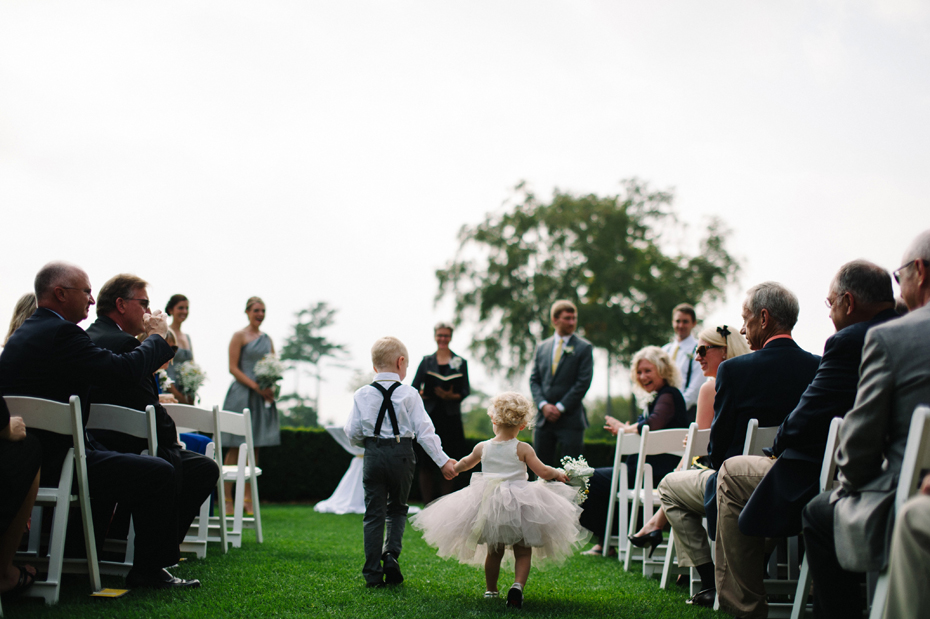 The flower girl wearing a tutu and the ring bearer wearing suspenders come down the aisle at a wedding ceremony held outside at The Inn at Stonecliffe on Mackinac Island by Detroit Wedding Photographer Heather Jowett.