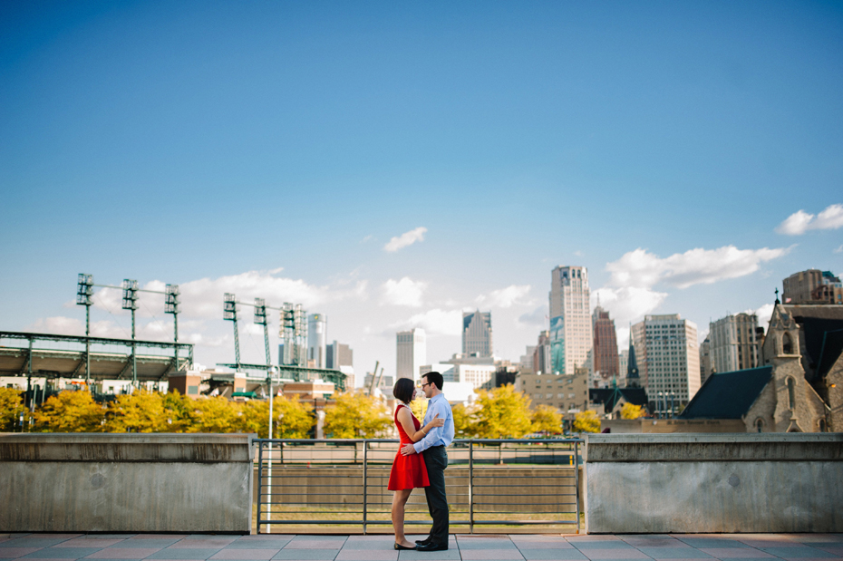 A couple poses for an engagement photo with Comerica Park and the Detroit Skyline in the background.