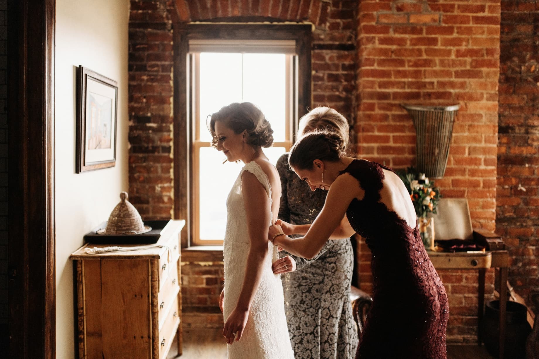 Bride helped into her dress by her mother and sister by Detroit wedding photographer Heather Jowett