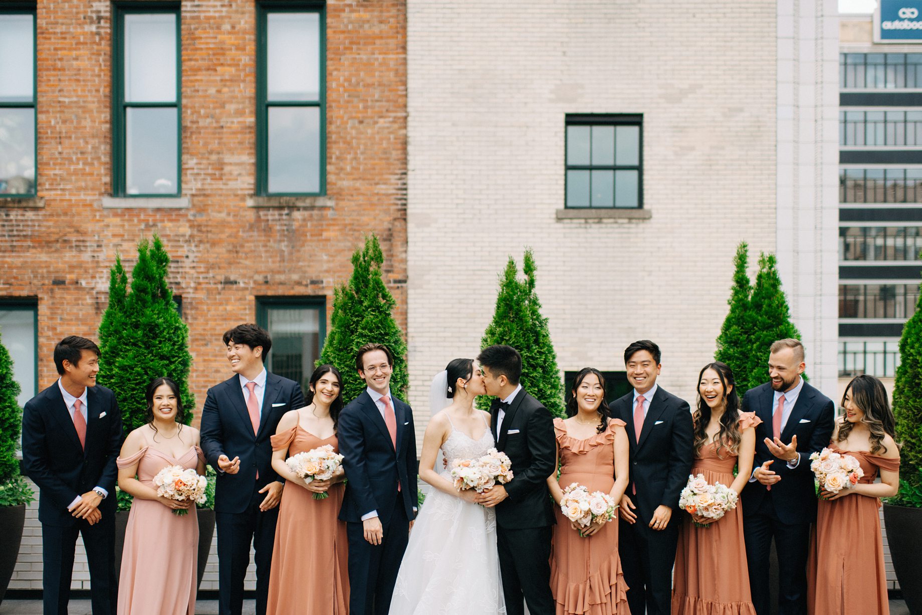 The full wedding party celebrates on the rooftop of the Shinola Hotel with the couple kissing in the center
