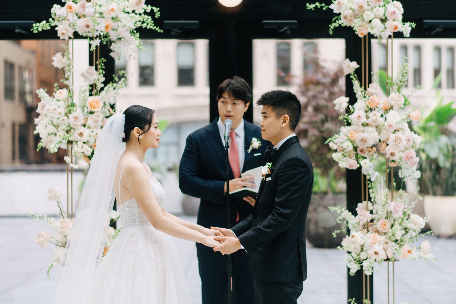 A close up of the bride and groom holding hands and exchanging vows beneath floral pillars in the Shinola Hotel atrium surrounded by soft light and downtown views in the birdy room