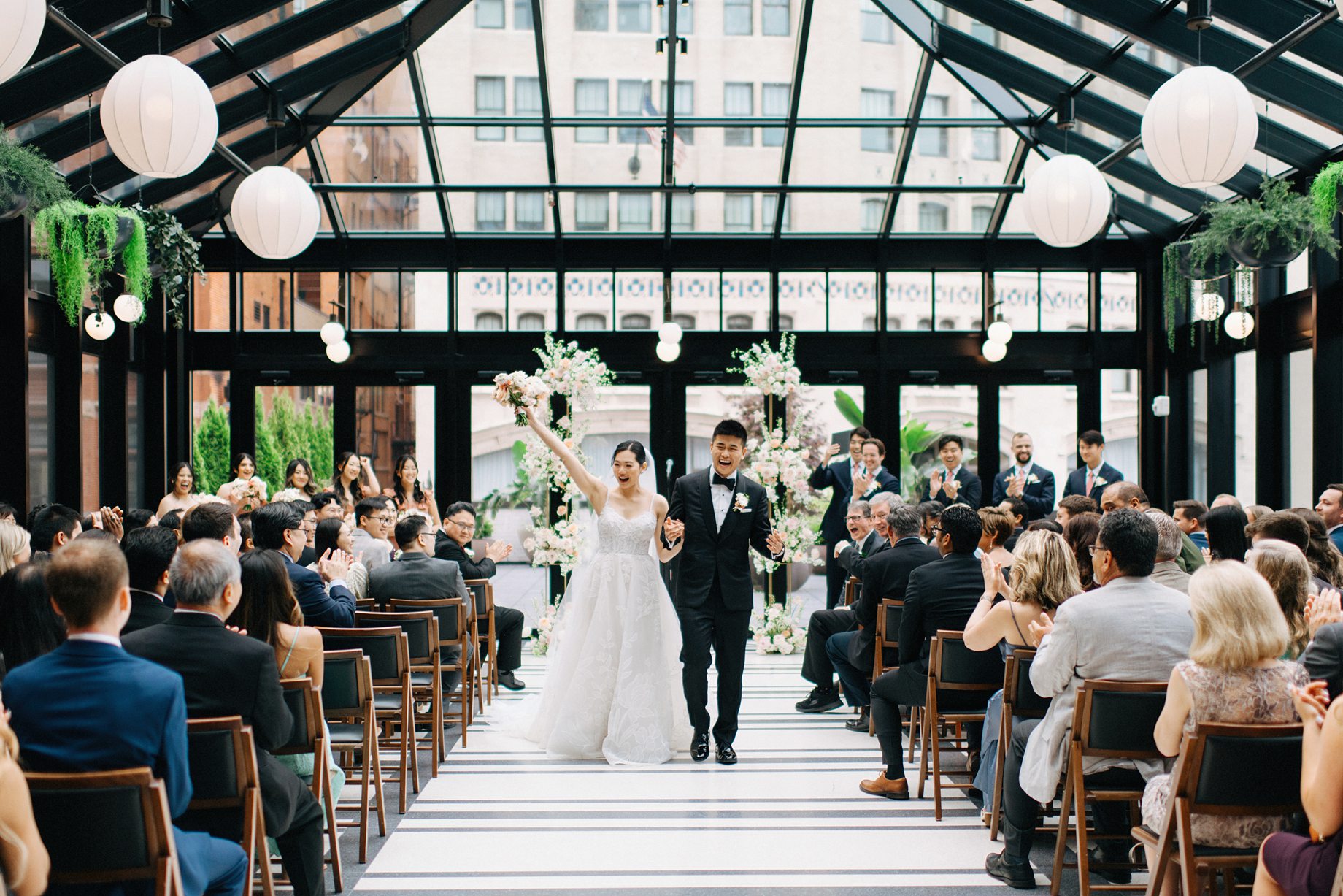 The bride and groom recess joyfully back down the aisle as newlyweds hands raised in celebration while guests cheer from their seats
