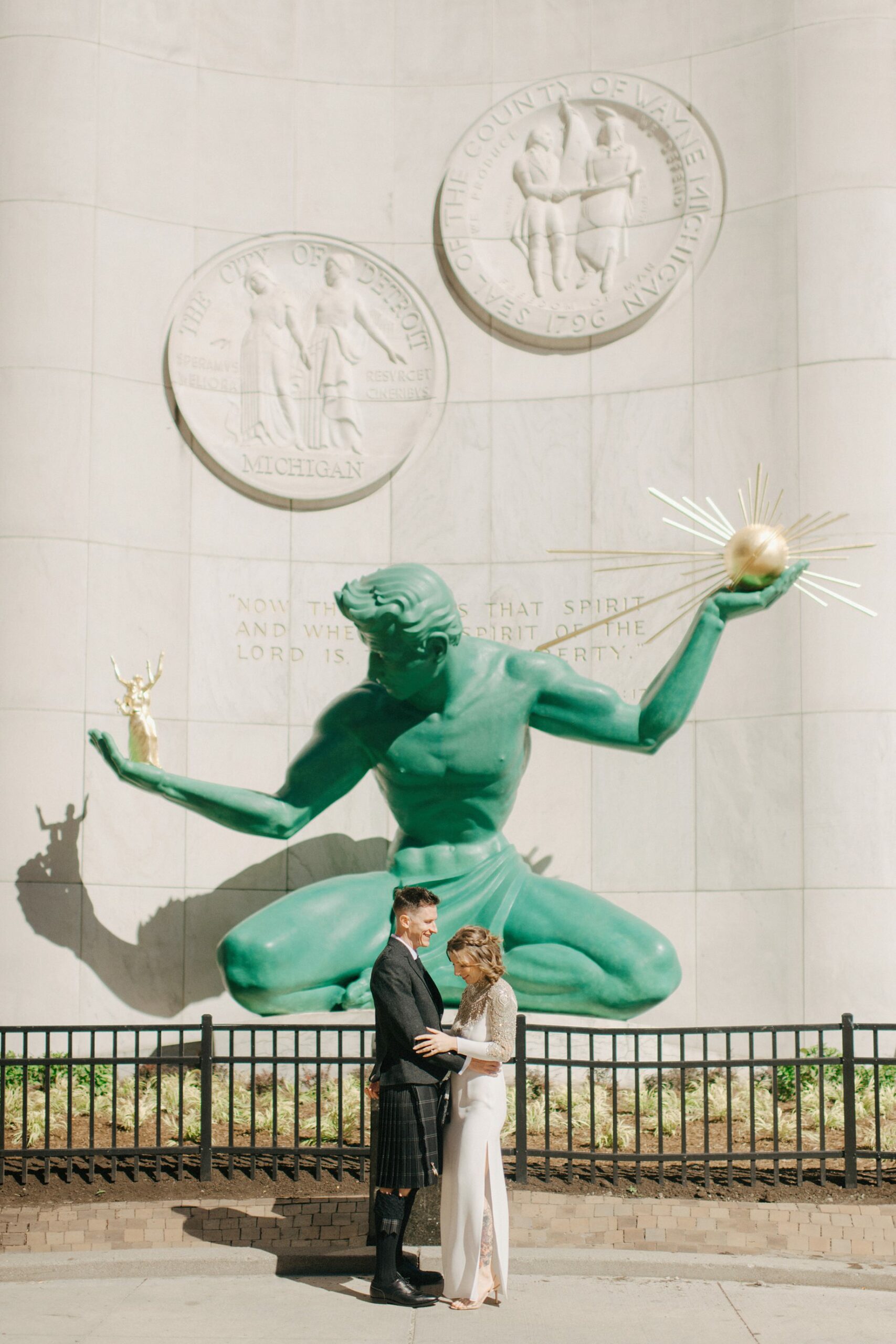 Bride and groom stand in front of the Spirit of Detroit statue in an iconic Detroit wedding photo at a landmark location near the Guardian Building