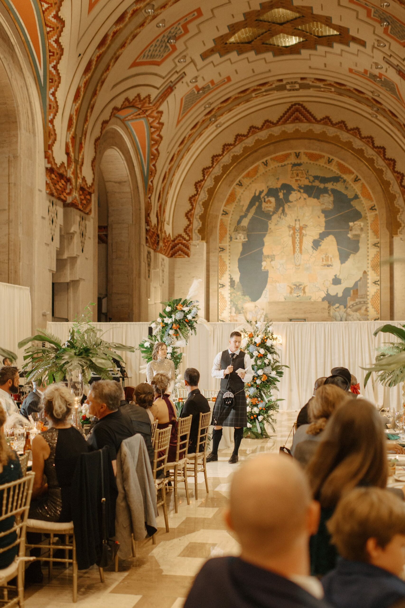 groom speaking during a wedding reception at the Guardian Building with guests in the foreground and the iconic Michigan Mural in the background