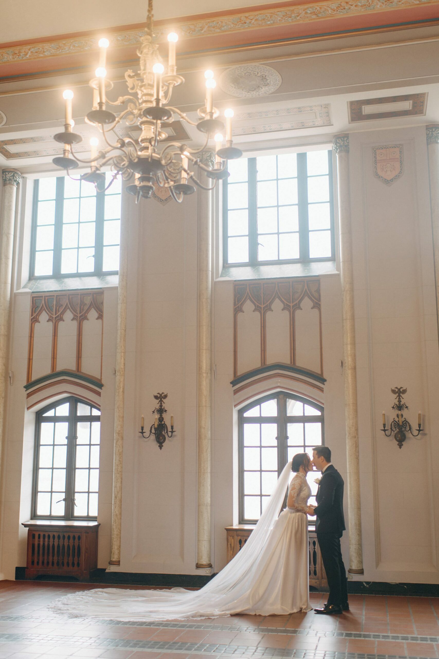 Wide shot of bride and groom kissing beneath a grand chandelier at their Detroit Masonic Temple wedding showcasing architectural drama and heartfelt connection