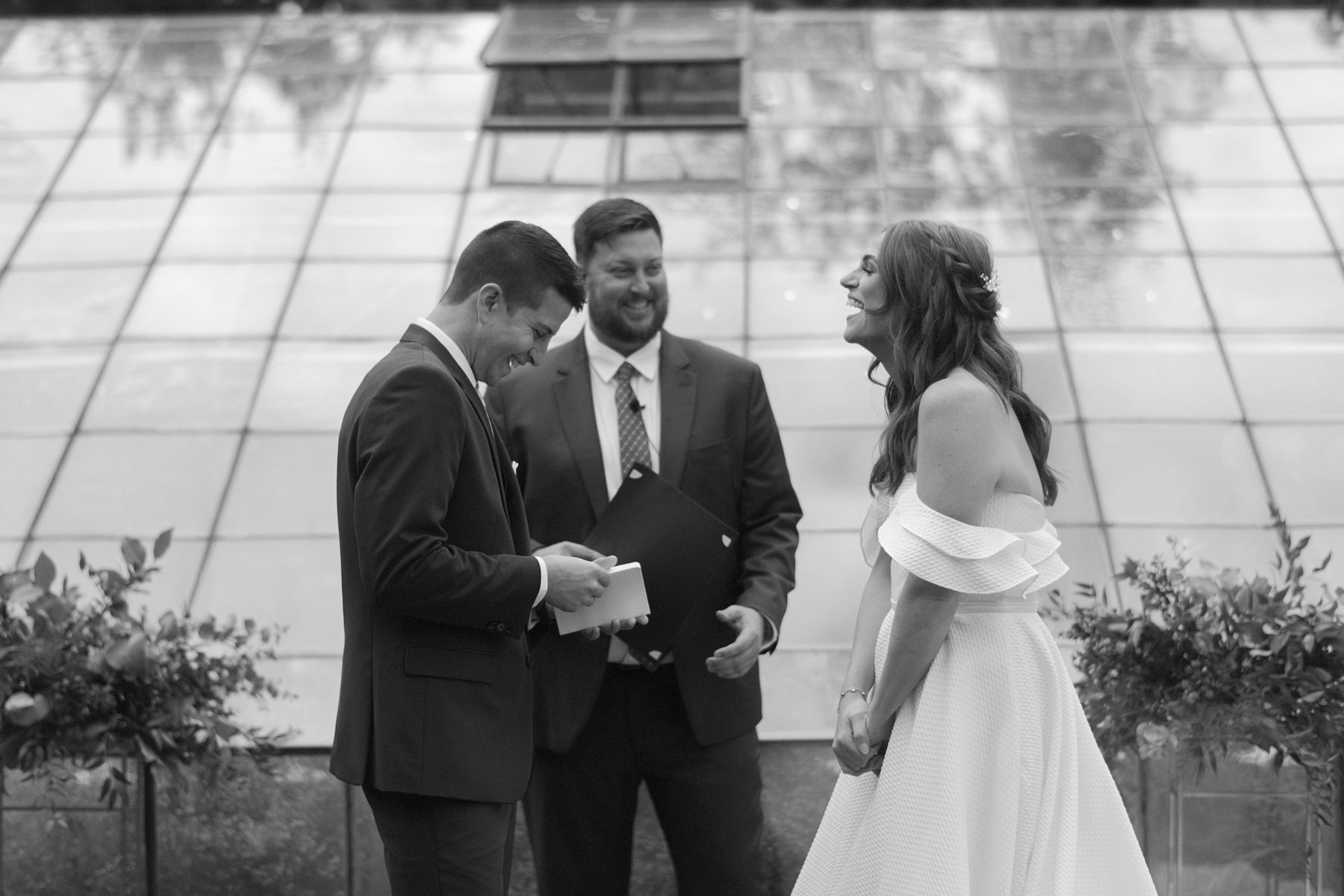 Bride and groom laugh while reading vows during ceremony at Glass House Community