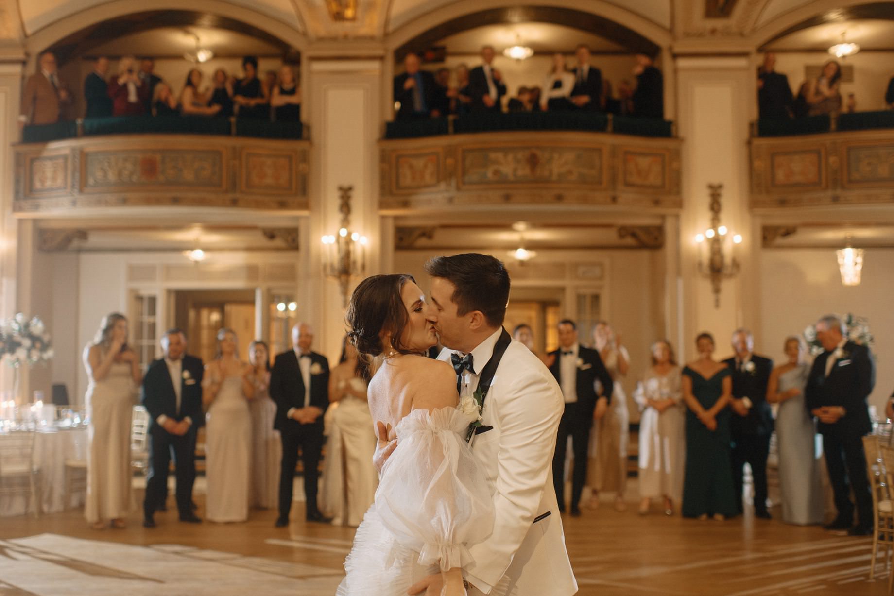 Groom kisses bride on the dance floor in the Crystal Ballroom at the Detroit Masonic Temple with friends and family looking on captured by luxury Detroit wedding photographer Heather Jowett