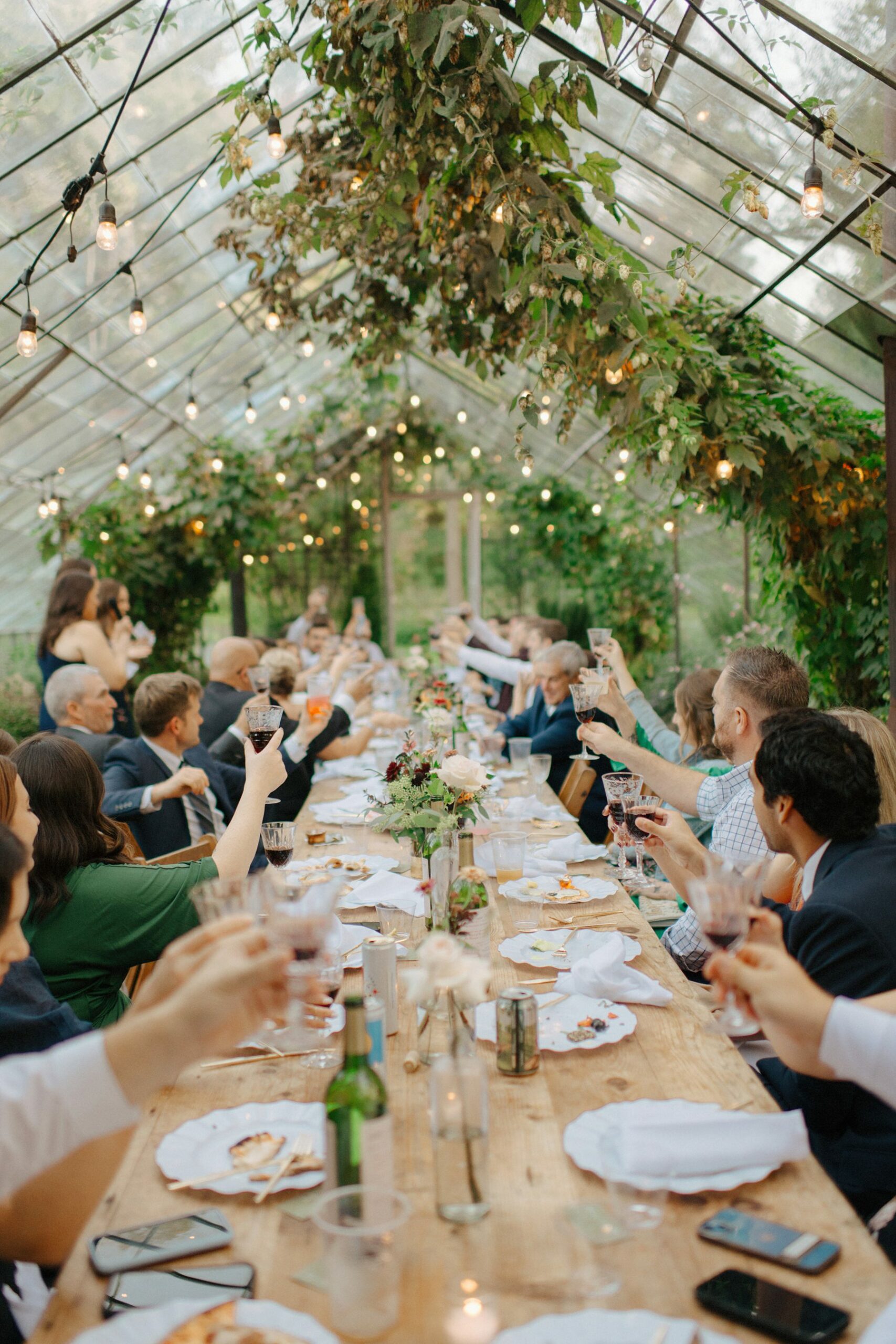 Guests clinking glasses across a long wooden dinner table framed by greenery and candlelight in the greenhouse at the Class House Community