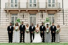 bride and groom pose with their wedding party in tuxedos and pale blue grey