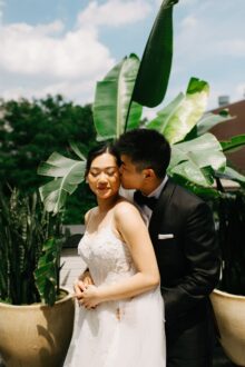The groom kisses the brides cheek in a romantic moment beneath tropical foliage on the Shinola Hotel rooftop