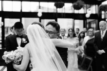 The bride hugs her father at the end of the aisle as the groom watches tenderly in the background
