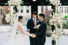 A close up of the bride and groom holding hands and exchanging vows beneath floral pillars in the Shinola Hotel atrium surrounded by soft light and downtown views in the birdy room