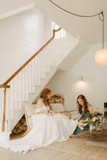 Bride sits on modern wooden bench while sister helps with her shoes