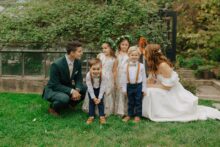 Bride and groom crouch to laugh with flower girls and ring bearers in front of greenhouse