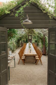 Long farm table reception inside a lush greenhouse with vintage wood chairs and string lights for an elegant garden wedding