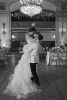 Black and white photo groom lifting bride beneath a grand chandelier during their first dance at the Detroit Masonic Temple an iconic moment captured by Detroit wedding photographer Heather Jowett